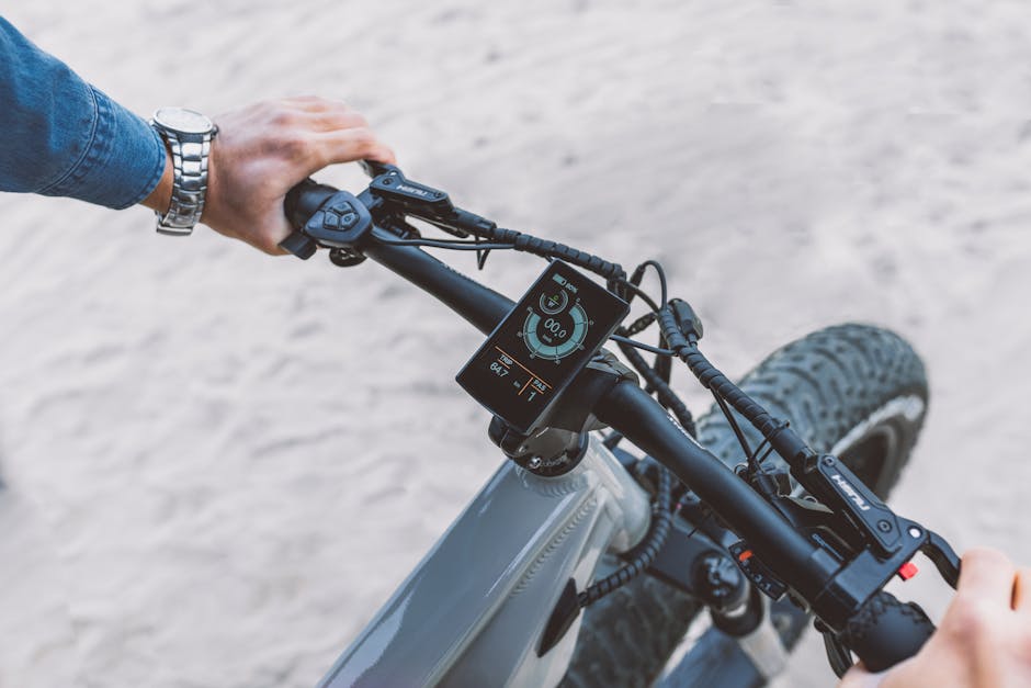 A modern electric bicycle handlebar with a touch screen display, featuring riding statistics on a sandy outdoor surface.