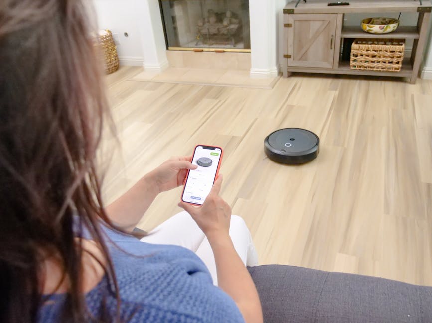 Overhead view of a woman controlling a robot vacuum using a smartphone app on a wooden floor.