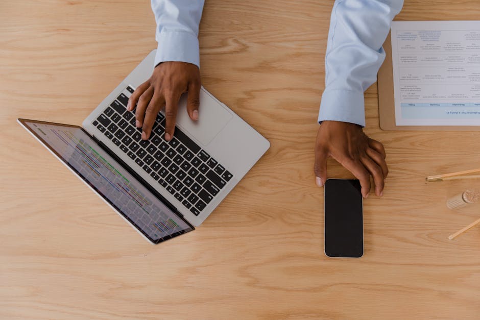 Top view of a workspace featuring hands using a laptop and smartphone on a wooden desk.