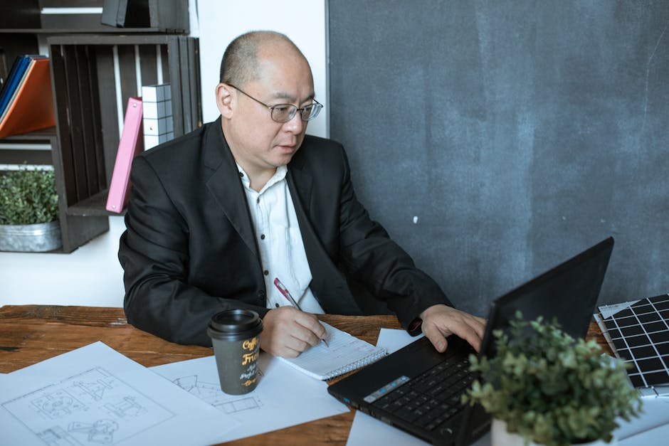 A focused businessman working on a laptop in his home office, taking notes and sipping coffee.