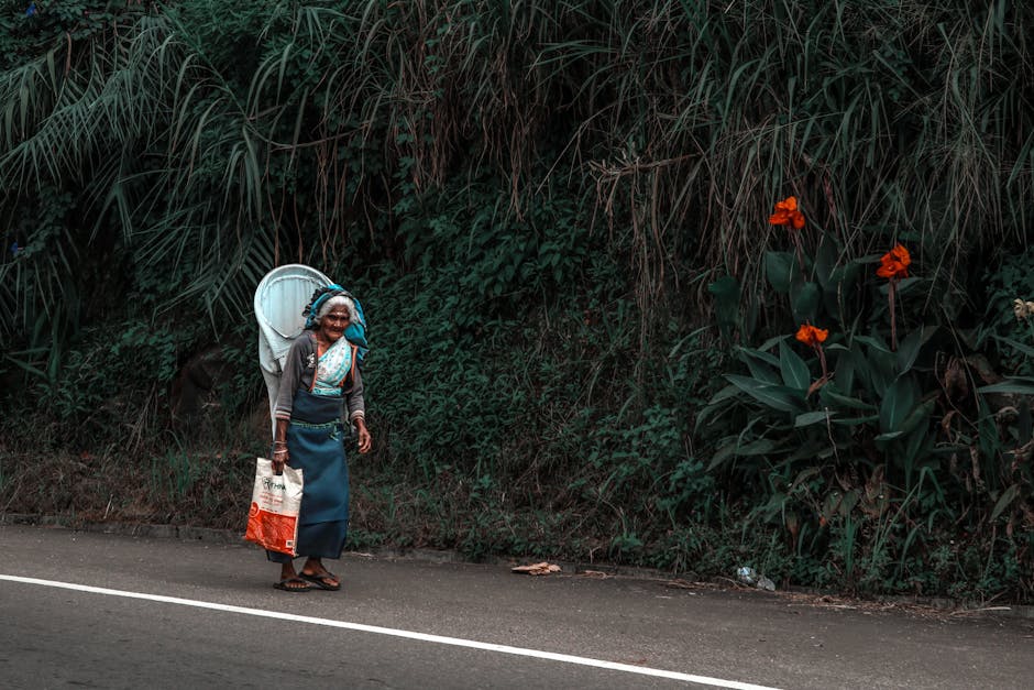 An elderly woman walking on a rural road beside lush greenery and vibrant flowers in Sri Lanka.