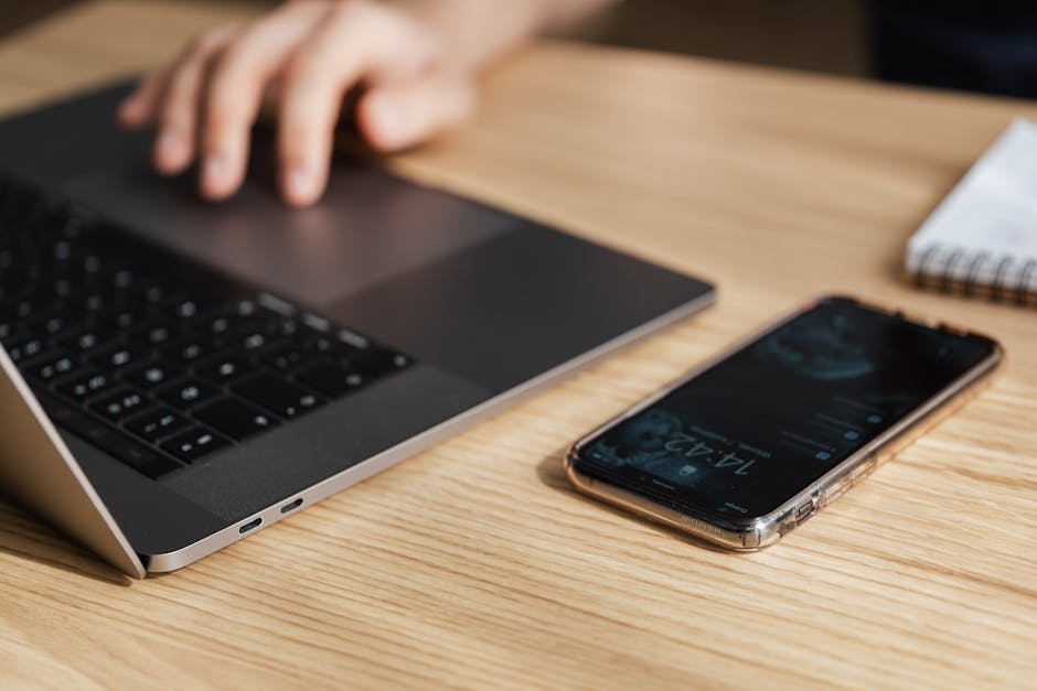 A hand on a laptop next to a smartphone on a wooden desk, symbolizing modern work life.