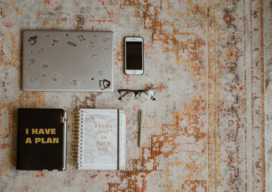 Flat lay of workspace items including laptop, phone, planners, and glasses on a patterned rug.
