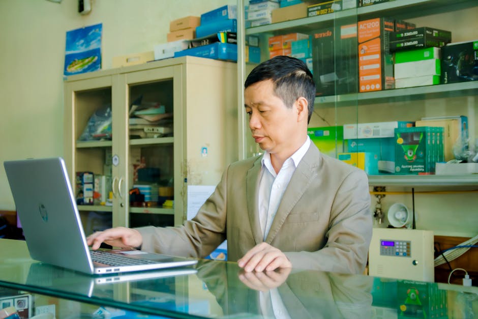 A man working on a laptop in an office surrounded by shelves of tech products.