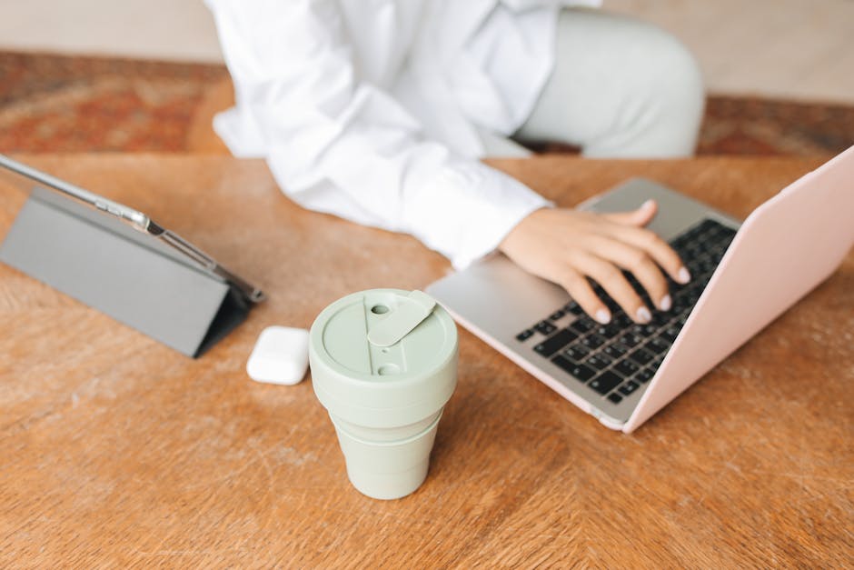 A person working remotely on a laptop with a tablet and reusable cup on a wooden table.