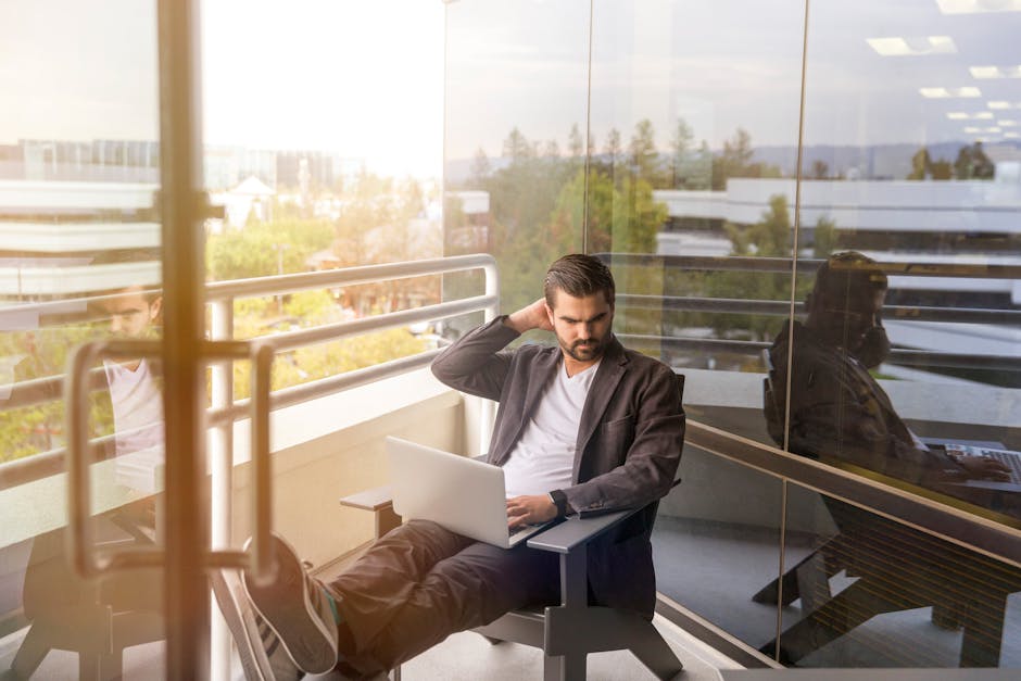 Man working remotely with laptop on office balcony, enjoying natural daylight and modern office view.
