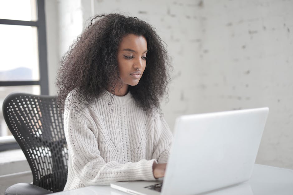 A young woman with curly hair working on a laptop in a bright, modern office setting.
