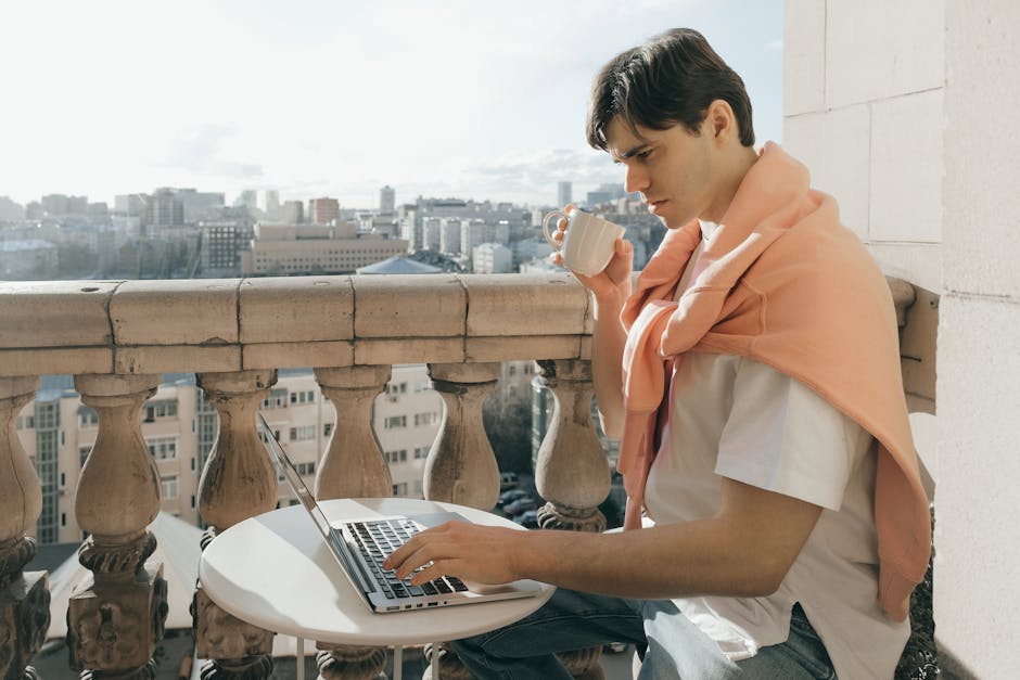 Man enjoys coffee while working on a laptop on a city balcony.