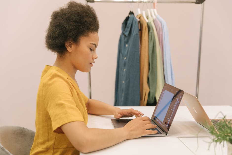 Young woman in fashion studio using laptop, focused on work with clothing rack in background.