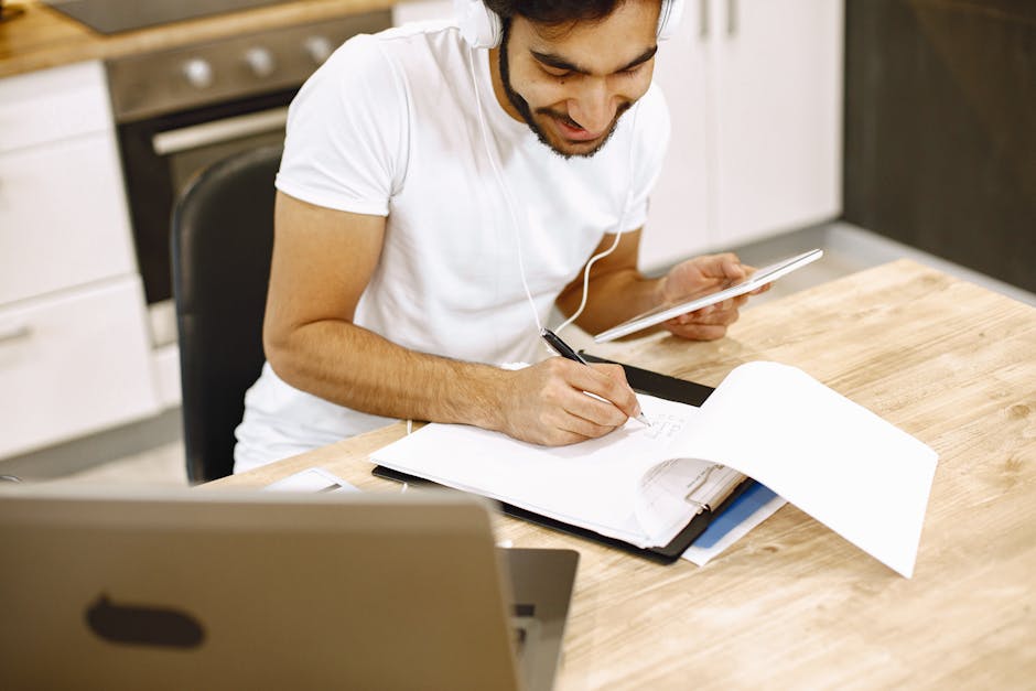 Adult man takes notes while using tablet and laptop in a modern kitchen setting.