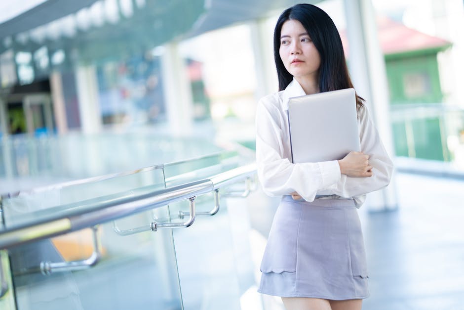 Young Asian businesswoman holding a laptop in a modern office environment, showcasing confidence and professionalism.