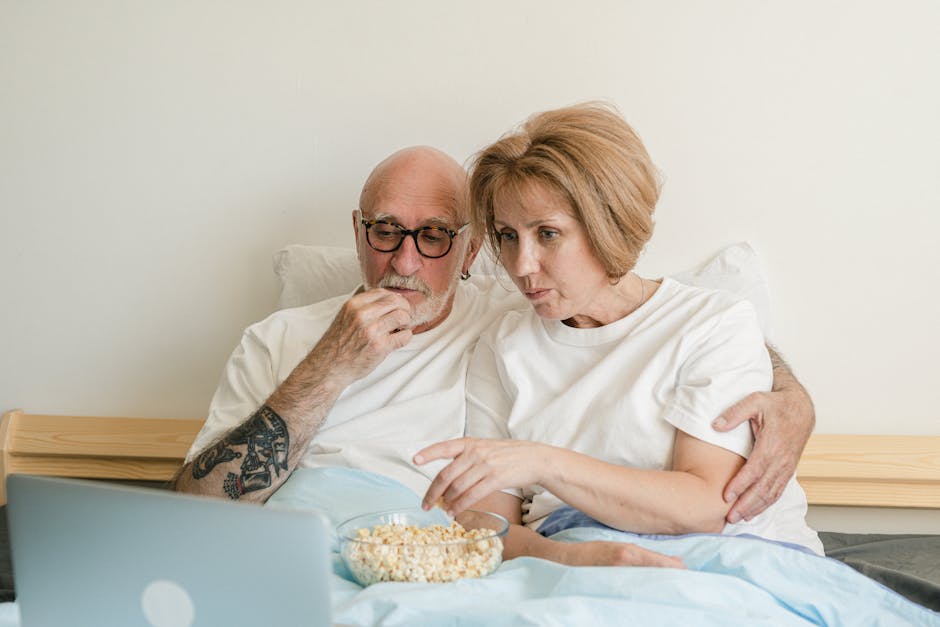 Senior couple bonding over movie night with popcorn and laptop in cozy home setting.