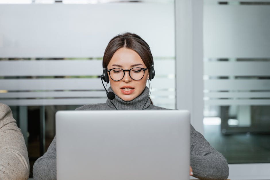 Focused woman with eyeglasses and headset working on laptop in a modern office environment.