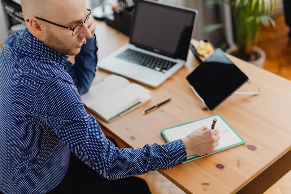 Adult man writing notes at a wooden desk with a laptop, phone, and plant indoors.